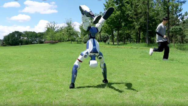 Ein Unitree R1 Roboter macht einen Handstand auf einer Wiese, während eine Person im Hintergrund joggt.
