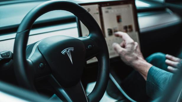 Das Lenkrad eines Tesla mit dem Tesla-Logo und ein Mann, der den Touchscreen bedient.