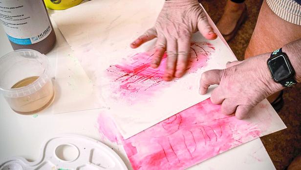 Eine ältere Frau malt mit den Fingern pinke Farbe auf Papier.
