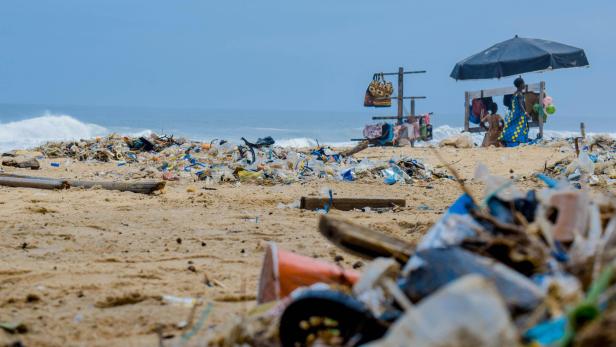 Ein Strand ist mit Müll übersät, im Hintergrund ein Verkaufsstand mit zwei Personen.