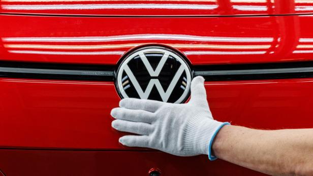 Nahaufnahme auf das VW Logo eines roten Fahrzeugs, daneben eine behandschuhte Hand.
