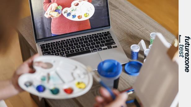 Eine Person hält eine Malerpalette, im Hintergrund eine Leinwand und ein Laptop, auf dem ebenfalls eine Malerpalette zu sehen ist.
