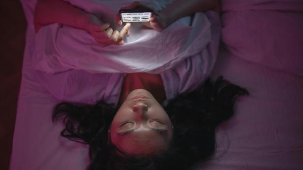 Eine Frau liegt auf ihrem Bett und scrollt auf ihrem Smartphone im Dunklen.