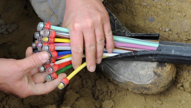 Zwei Hände halten bunte Einzelstränge eines Glasfaserkabels in einer sandigen Grube.