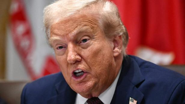 Donald Trump spricht ernsthaft, trägt einen dunklen Anzug mit US-Flaggen-Pin und sitzt vor roten Fahnen.