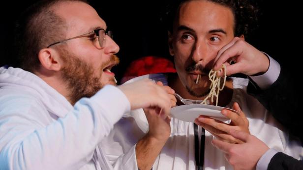 Zwei Männer essen gemeinsam Spaghetti von einem Teller, während mehrere Hände ihnen helfen.