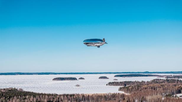 Ein silbernes Luftschiff fliegt über eine verschneite Wald- und Seenlandschaft.