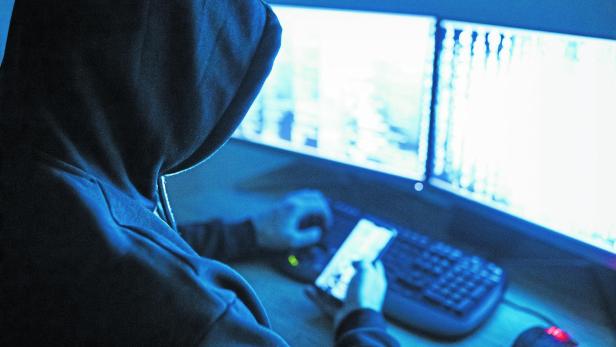 Hacker attacking internet Eine Person mit Kapuze sitzt vor zwei hellen Monitoren und hält ein Smartphone in der Hand.