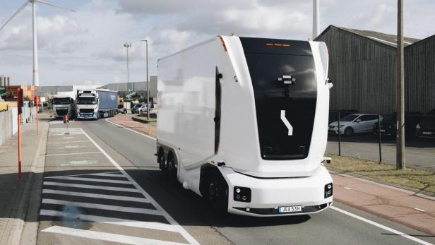 Ein weißer, autonomer Lkw mit schwarzer Front fährt auf einer Straße mit Windrädern im Hintergrund.
