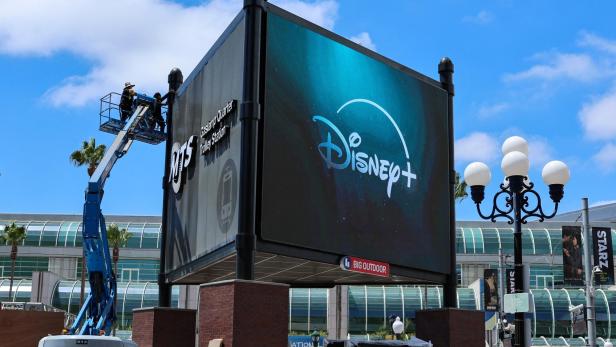 Zwei Personen arbeiten auf einer Hebebühne an einer großen Werbetafel mit Disney+ Logo an einer Straßenkreuzung.