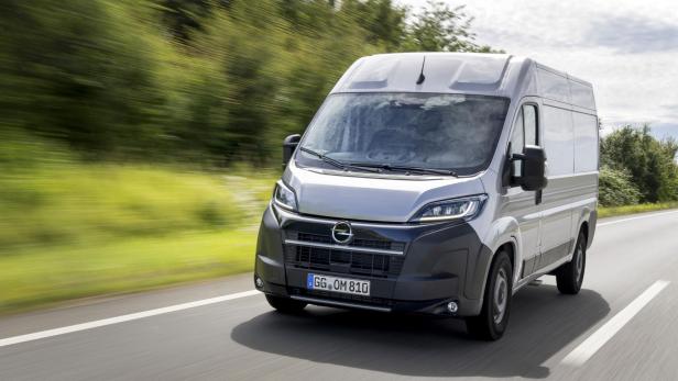 Ein Kastenwagen fährt auf einer Straße, im Hintergrund sieht man grüne Bäume.
