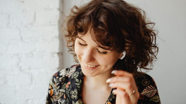 Eine junge Frau mit lockigen Haaren lächelt und trägt kabellose Ohrhörer.