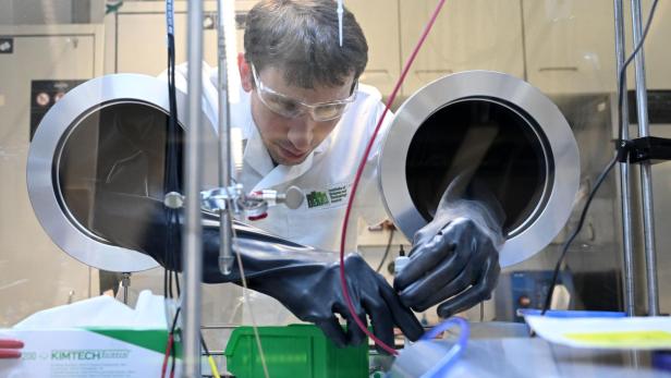 Ein Mann arbeitet mit Schutzhandschuhen in einer geschlossenen Laborapparatur an technischen Geräten.