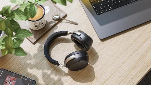Der Sennheiser HDB 630 Kopfhörer liegt auf einem Schreibtisch neben einem Laptop, einer Tasse Kaffee und einem Smartphone mit geöffneter Musik-App.