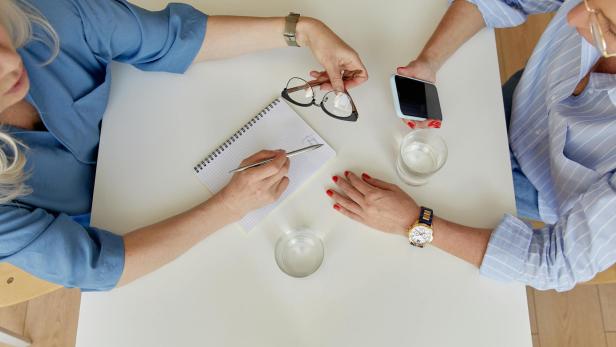 Zwei Personen sitzen an einem Tisch und unterhalten sich, eine Person hält ein Smartphone, die andere einen stift