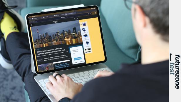 Eine Person sitzt auf einem Sofa und nutzt ein Tablet mit Tastatur, auf dem eine Technik-Webseite geöffnet ist.