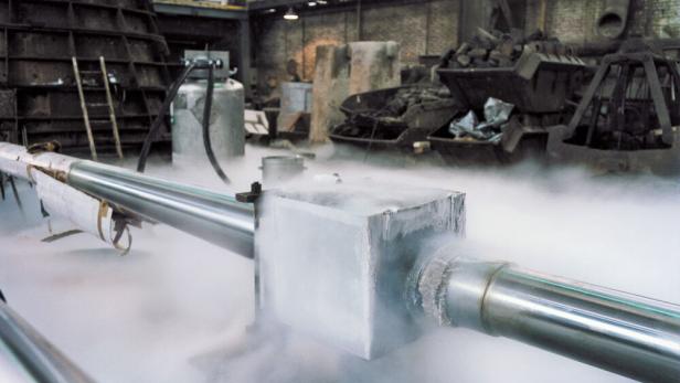 Metallrohre mit frostbedecktem Verbindungsstück, aus dem kalter Dampf entweicht, in einer Industriehalle.