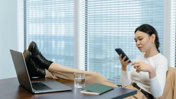 Eine Frau, die ihre Füße auf dem Schreibtisch liegen hat, hält ein Smartphone in der Hand 