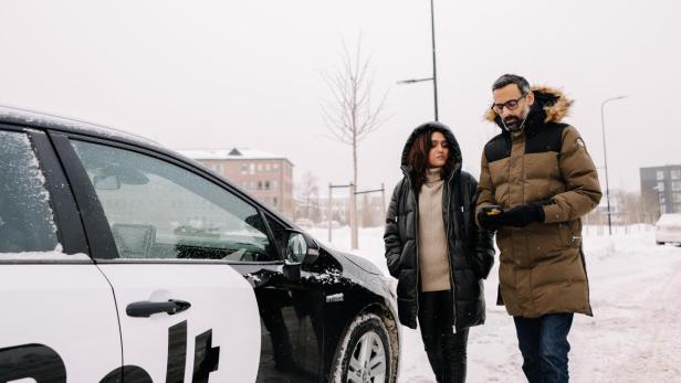 Zwei Personen gehen im Winter neben einem Bolt-Auto auf verschneiter Straße.