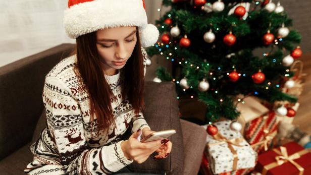 Frau mit weihnachtsmütze neben Christbaum und Geschenken schaut in ihr Smartphone