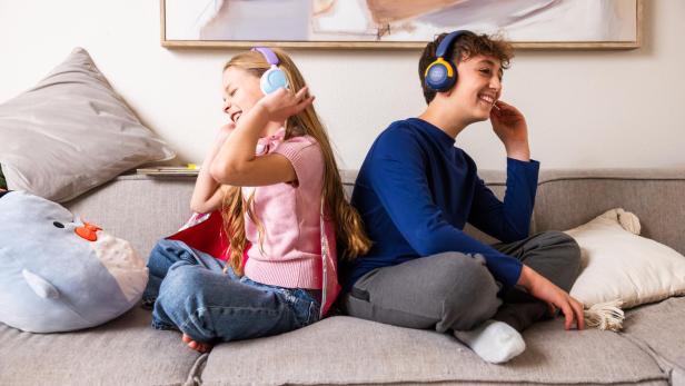 Zwei Kinder sitzen lachend auf einem Sofa und tragen die myFirst CareBuds Max Kopfhörer in verschiedenen Farben.