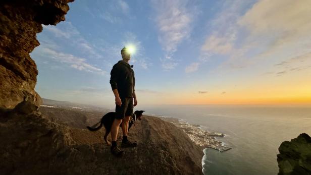 Mann mit Stirnlampe und Hund steht auf einer Klippe in der Dämmerung