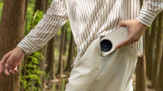 Eine Person steckt ein Smartphone mit großem Kameramodul in eine Hosentasche, während sie durch einen Wald geht.