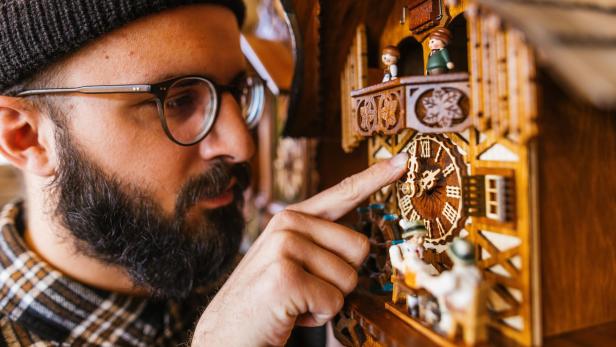 Ein Mann mit Brille und Mütze stellt die Zeit an einer Kuckucksuhr.