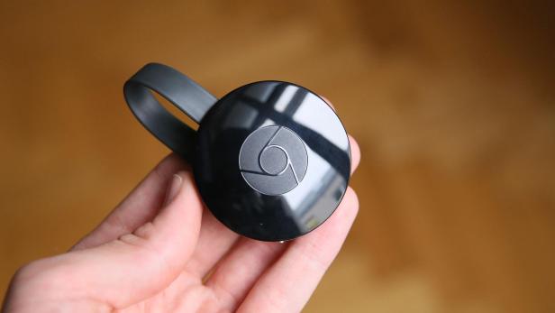 Eine Hand hält einen schwarzen Google Chromecast vor unscharfem Hintergrund.