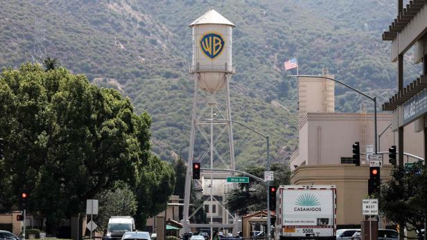 Großer Wasserturm mit WB-Logo vor bewaldetem Hügel, Autos und Fußgänger an einer Kreuzung.