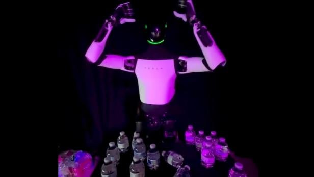 Der Tesla-Roboter Optimus steht auf einem Event in Miami hinter einem Tisch mit mehreren Wasserflaschen und hebt beide Arme.