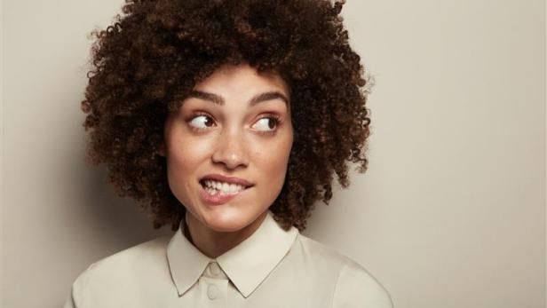 Eine junge Frau mit lockigem Haar schaut zur Seite und beißt sich nachdenklich auf die Lippe, passend zum Thema Zahnpflege.