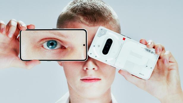 Eine Person hält ein Smartphone mit einem vergrößerten Auge auf dem Display vor ihr Gesicht und daneben ein weiteres Smartphone mit sichtbarer Rückseite.