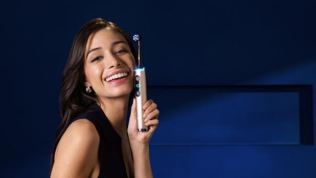 Eine Frau hält lächelnd die Oral-B iO10 Gold Special Edition Zahnbürste vor einem dunkelblauen Hintergrund.