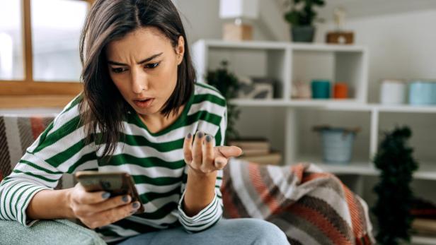 Eine junge Frau mit grün-weiß gestreiftem Pullover schaut verärgert auf ihr Smartphone und gestikuliert mit der Hand.
