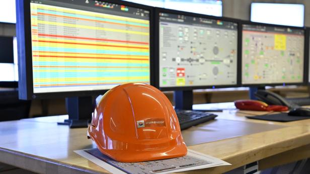 Ein orangefarbener Schutzhelm liegt auf einem Schreibtisch vor mehreren Monitoren mit technischen Übersichten und Tabellen.