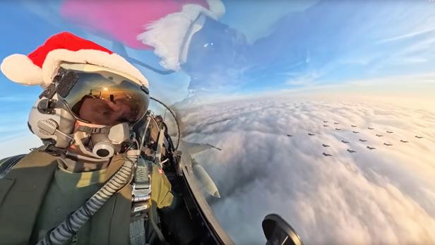 Pilot mit Weihnachtsmütze im Cockpit, dahinter Kampfjets in Weihnachtsbaum-Formation am Himmel.