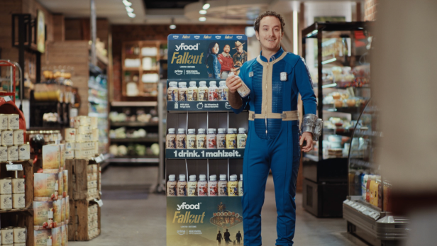 Eine Person in einem blauen Fallout-Anzug steht vor einem yfood-Promostand mit Fallout-Getränken in einem Supermarkt.