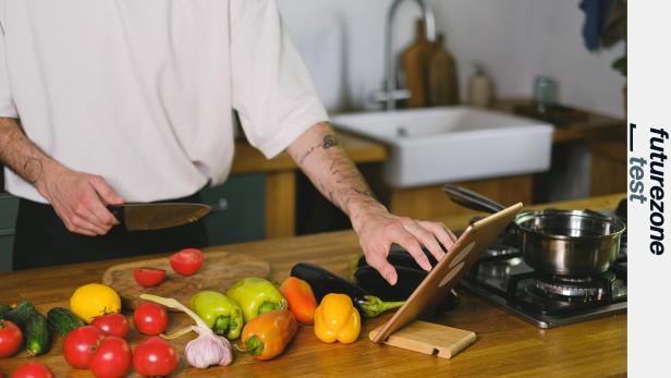 ein Mann in der Küche hält ein Messer und tippt mit der anderen Hand auf ein Tablet, vor ihm frisches Gemüse und ein Kochfeld