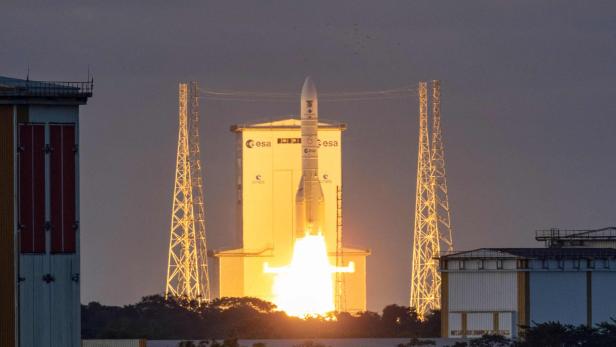 Eine Rakete startet mit heller Flamme von einer Rampe, umgeben von Gebäuden und Türmen bei Dämmerung.