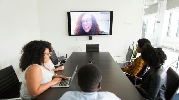 Vier Personen sitzen im Konferenzraum und nehmen an einem Videocall mit einer Person am Bildschirm teil.