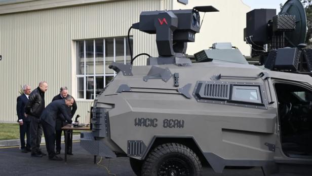 Gepanzertes Fahrzeug mit Laserwaffe Magic Beam, daneben mehrere Männer im Anzug und Lederjacke.