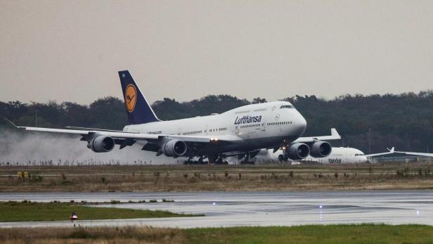 Ein großes Passagierflugzeug mit Lufthansa-Logo landet auf einer nassen Landebahn, im Hintergrund steht ein weiteres Flugzeug.
