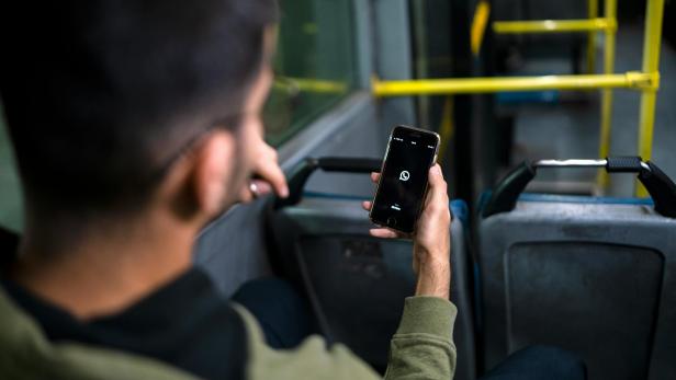Ein Mann sitzt in öffentlichem Verkehrsmittel mit Smartphone, auf dem WhatsApp geöffnet wird 