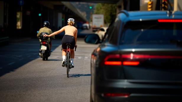 Radfahrerin mit Helm zeigt Handzeichen, hinter ihr Auto und Roller im Straßenverkehr.