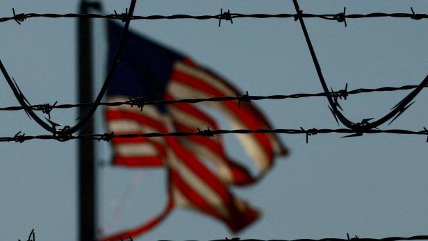 Stacheldraht im Vordergrund, im Hintergrund weht unscharf eine amerikanische Flagge im Wind.
