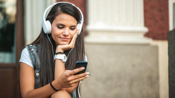 Junge Frau mit langen dunklen Haaren hört mit weißen Kopfhörern Musik und schaut auf ihr Smartphone.