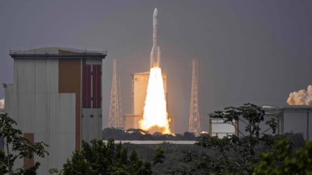 Eine Rakete startet mit heller Flamme und Rauch von einer Abschussrampe, umgeben von Gebäuden und Bäumen.