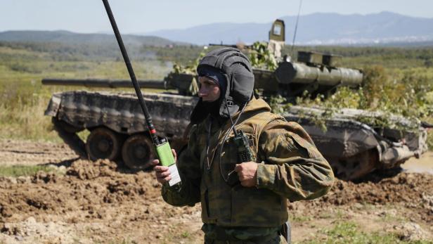 Ein Soldat in Tarnkleidung mit Funkgerät steht vor einem Panzer auf schlammigem Gelände.