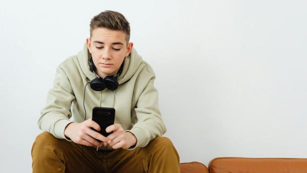 Junger Mann sitzt mit Kopfhörern um den Hals und blickt auf sein Smartphone